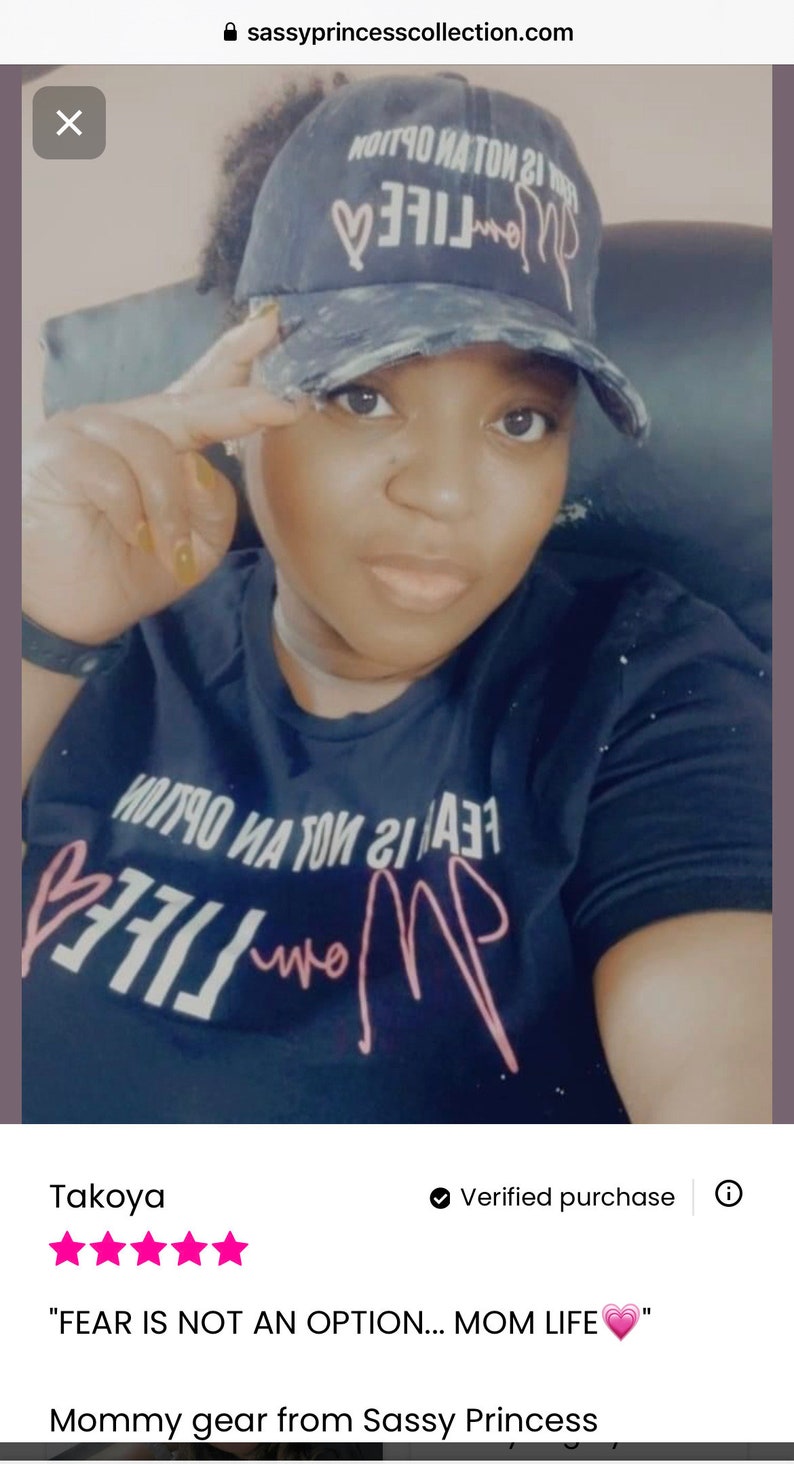 May include: A black t-shirt with the text "FEAR IS NOT AN OPTION... MOM LIFE" in pink lettering with a pink heart. The person wearing the shirt is also wearing a black and white baseball cap with the text "MOM LIFE" in pink lettering with a pink heart.