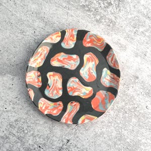 May include: A round, black ceramic plate with a colorful pattern. The pattern features abstract shapes in shades of red, orange, blue, and yellow. The plate has a glossy finish and is suitable for serving food.