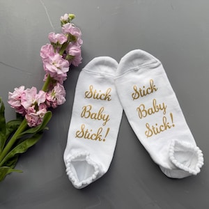 Op de afbeelding: Een paar witte babysokken met de gouden tekst "Stick Baby Stick!". De sokken hebben een gerimpelde rand. Een takje roze bloemen ligt naast de sokken op een grijze ondergrond.