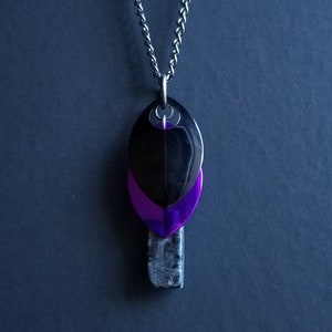 May include: A silver chain necklace with a pendant featuring three overlapping teardrop shapes. The top shape is black, the middle shape is purple, and the bottom shape is a gray stone.
