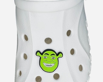 Pode incluir: Um sapato branco com um encanto de personagem Shrek verde e preto. O encanto é uma imagem de desenho animado do rosto de Shrek, com uma tez verde, sobrancelhas pretas e um sorriso largo. O sapato tem vários orifícios de ventilação.