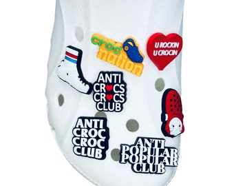 May include: White Crocs shoe adorned with various colorful charms. Charms include a sneaker, a red heart with text, and several charms with text like "Croc Nation" and "Anti Crocs Club".
