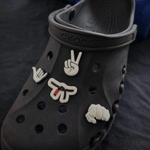 Peut inclure: Sabot Crocs noir orné de breloques blanches et noires. Les breloques comprennent un geste de la main, un signe de paix, une ceinture de karaté et un poing. Le mot "Crocs" est visible sur la bride.