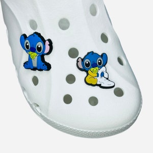 Può includere: Scarpa Crocs bianca con due ciondoli Stitch blu e neri. Un ciondolo mostra Stitch seduto con un ciuccio, l'altro Stitch con un ciuccio e una coperta. I ciondoli sono colorati e aggiungono un tocco giocoso.
