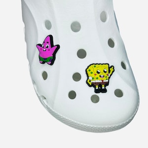 Op de afbeelding: Witte schoen met twee cartoonbedels. De ene bedel is een roze zeester met een zwarte omtrek en een groene korte broek. De andere bedel is een geel vierkant karakter met een zwarte omtrek, een bruine broek en een rode stropdas.