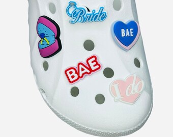 May include: White shoe with decorative charms. Charms include a blue "Bride" design, a pink and blue heart with "BAE", a ring in a box, a red "BAE" charm, and a pink heart with "I do".