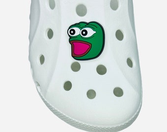 May include: A white clog shoe with a green and pink cartoon frog charm attached. The charm features a green frog face with large eyes and an open mouth, set against a white background.