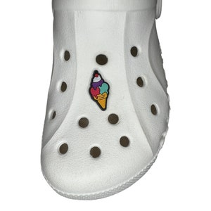 Puede incluir: Un zapato blanco con un charm de helado. El charm presenta un cono de helado colorido con bolas rojas, moradas, turquesas y amarillas. Una pequeña cereza roja encima.