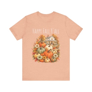 May include: A heather peach short-sleeve t-shirt featuring a graphic print of a white farmhouse surrounded by various pumpkins, gourds, and autumn flowers. The text "HAPPY FALL Y'ALL" is printed in a white, thin, sans-serif font above the illustration.