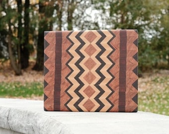 Op de afbeelding: Een vierkant houten snijplank met een geometrisch patroon van bruin, zwart en licht hout. De plank ligt op een grijs stenen oppervlak.