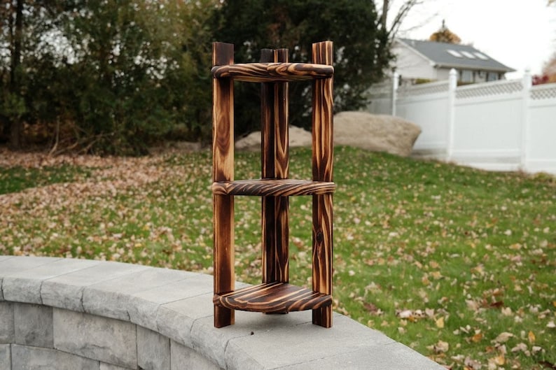 Puede incluir: Una estanter&iacute;a de esquina de tres niveles hecha de madera con un acabado quemado. La estanter&iacute;a est&aacute; sobre una pared de piedra.