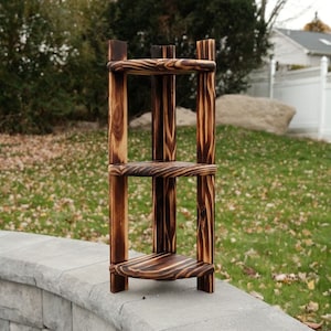 Puede incluir: Una estanter&iacute;a de esquina de tres niveles hecha de madera con un acabado quemado. La estanter&iacute;a est&aacute; hecha de gruesas tablas de madera y tiene un aspecto r&uacute;stico.
