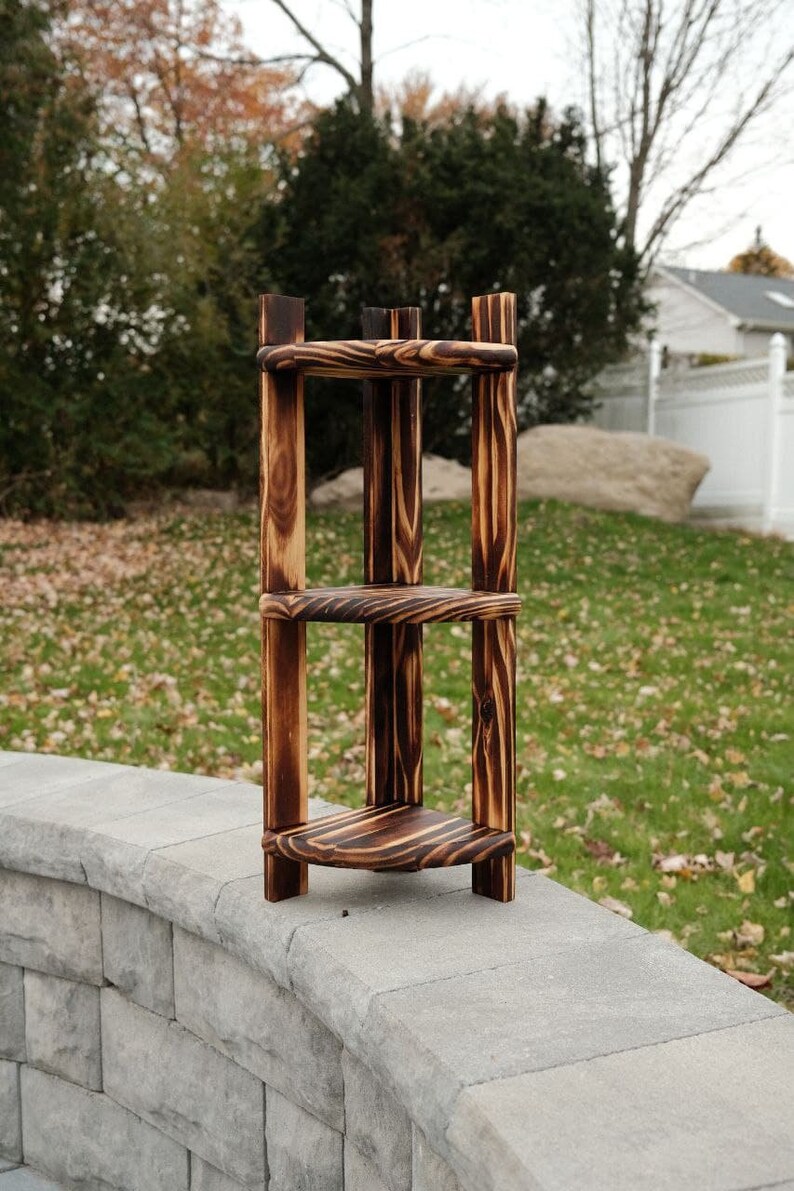 Puede incluir: Una estanter&iacute;a de esquina de tres niveles hecha de madera con un acabado quemado. La estanter&iacute;a est&aacute; hecha de gruesas tablas de madera y tiene un aspecto r&uacute;stico.