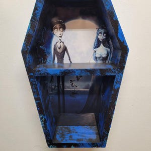 May include: A coffin-shaped shelf painted blue with black accents. Inside, it features a scene from a movie, with characters and a backdrop. The shelf is designed to be wall-mounted, adding a unique decorative element.