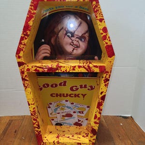 May include: A yellow coffin-shaped display box with red accents and blood splatter. The box features a Chucky doll with red hair and stitched facial features. The box has the words "Good Guy Chucky" on the front.