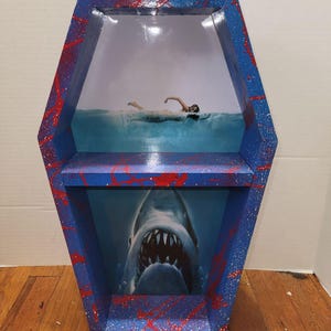 Puede incluir: Estantería con forma de ataúd con exterior azul, pintura salpicada roja y un estante. El interior presenta una imagen de tiburón debajo y un nadador arriba. Una estantería decorativa única.