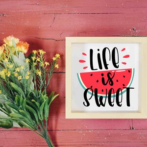 Puede incluir: Un cartel enmarcado en blanco con una rodaja de sand&iacute;a roja y el texto "Life is Sweet" en letra cursiva negra. El cartel est&aacute; sobre un fondo de madera roja con un ramo de flores amarillas.