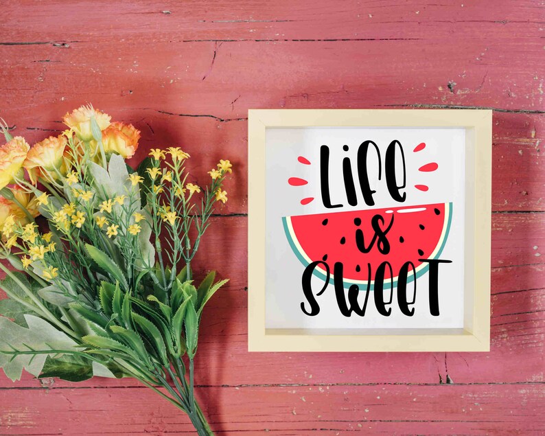 Puede incluir: Un cartel enmarcado en blanco con una rodaja de sand&iacute;a roja y el texto "Life is Sweet" en letra cursiva negra. El cartel est&aacute; sobre un fondo de madera roja con un ramo de flores amarillas.
