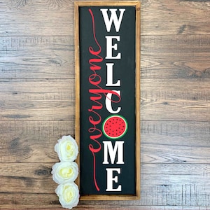 Puede incluir: Un letrero de madera negro con letras blancas que dice "Bienvenido a todos" con un gr&aacute;fico de sand&iacute;a roja en el centro.