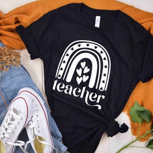 Może przedstawiać: Czarny T-shirt z białym wzorem tęczy i napisem "teacher" pod spodem. Tęcza ma małe serduszka wzdłuż łuku i większe serduszka w środku.