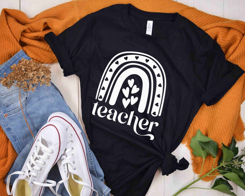 Może przedstawiać: Czarny T-shirt z białym wzorem tęczy i napisem "teacher" pod spodem. Tęcza ma małe serduszka wzdłuż łuku i większe serduszka w środku.