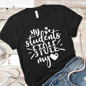Może przedstawiać: Czarny T-shirt z białym tekstem, kt&oacute;ry brzmi "Moi uczniowie ukradli mi serce" z małymi sercami wok&oacute;ł tekstu.