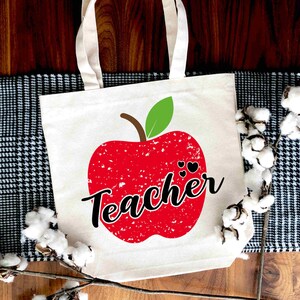 Może przedstawiać: Biała torba z pł&oacute;tna z nadrukiem czerwonego jabłka i napisem "Teacher" czarnym pismem. Jabłko ma fakturę postarzaną i dwa małe czarne serduszka.