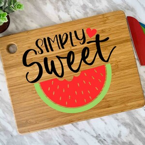 Puede incluir: Una tabla de cortar de madera con un dise&ntilde;o de sand&iacute;a y el texto "SIMPLY Sweet" con un coraz&oacute;n rojo.