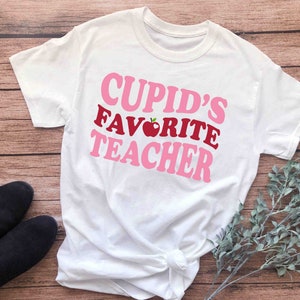 Może przedstawiać: Biała koszulka z r&oacute;żowym i czerwonym napisem "Cupid's Favorite Teacher" z grafiką czerwonego jabłka.