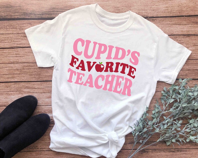 Może przedstawiać: Biała koszulka z r&oacute;żowym i czerwonym napisem "Cupid's Favorite Teacher" z grafiką czerwonego jabłka.
