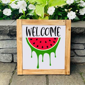 Puede incluir: Un letrero de madera con la palabra "WELCOME" en letras negras y una sand&iacute;a roja con c&aacute;scara verde y semillas negras que gotean jugo verde.