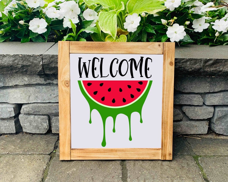 Puede incluir: Un letrero de madera con la palabra "WELCOME" en letras negras y una sand&iacute;a roja con c&aacute;scara verde y semillas negras que gotean jugo verde.