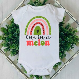 Puede incluir: Un body blanco para beb&eacute; con un arco&iacute;ris de sand&iacute;a y el texto "one in a melon" en rosa y verde.