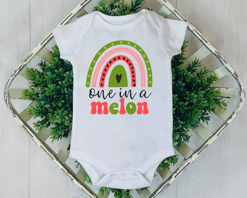 Puede incluir: Un body blanco para beb&eacute; con un arco&iacute;ris de sand&iacute;a y el texto "one in a melon" en rosa y verde.