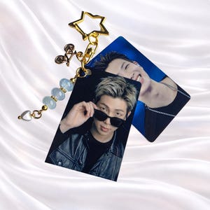 May include: A gold-toned star-shaped keychain featuring a small bicycle charm, three light blue beads, and a heart charm. Attached are two rectangular photo cards showing a person with blonde-streaked hair wearing sunglasses and a black jacket.