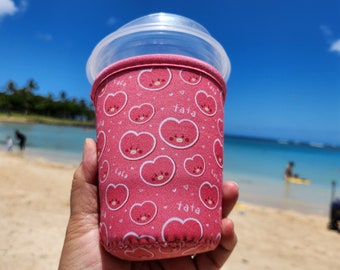 May include: Pink neoprene cup holder with a repeating pattern of pink hearts and the word "tata" printed on it.