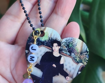 May include: A heart-shaped pendant necklace with a black and white photo of a person. The pendant has a gold chain and a purple flower charm with the letters "JM" on it.