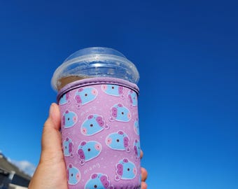 May include: A pink and blue insulated cup sleeve with a cartoon character pattern. The sleeve is made of neoprene and has a zipper closure. The sleeve is being held by a hand against a blue sky background.