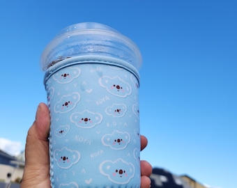 May include: A light blue drink sleeve featuring a pattern of white clouds and koala faces with the word "KOYA" printed on it. The sleeve is wrapped around a clear plastic cup with a dome lid, against a bright blue sky.