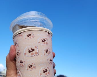 May include: A clear plastic cup with a lid, held in a hand, is encased in a beige insulated sleeve. The sleeve is patterned with cartoon bear faces. The background is a bright blue sky.