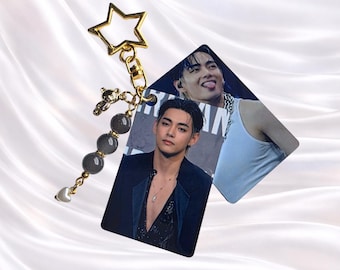 May include: A gold-toned star-shaped keychain featuring a small bear charm, three round grey beads, and a white heart charm. Attached are two square photo cards showing a person with dark hair in a dark blazer and a white tank top.