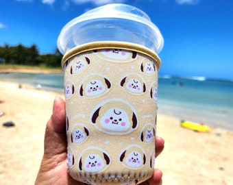 May include: A beige and brown insulated cup sleeve with a repeating pattern of cartoon dogs with pink cheeks. The sleeve is on a clear plastic cup.