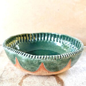 May include: A round ceramic bowl featuring a glossy dark green interior and a textured, fluted rim. The exterior displays a scalloped pattern where the green glaze meets the exposed, unglazed tan clay base, creating a distinct contrast.