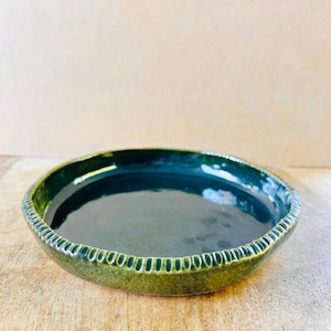 May include: A round ceramic dish with a glossy dark forest green glaze on the interior and a textured, ribbed edge along the outer rim. The dish features a deep basin and a slightly uneven, hand-crafted appearance with a reflective surface finish.