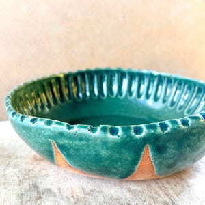 May include: A round ceramic bowl with a glossy deep green glaze on the interior and upper rim. The exterior features a textured, fluted edge and exposed terracotta-coloured clay at the base, revealing triangular patterns where the glaze ends.