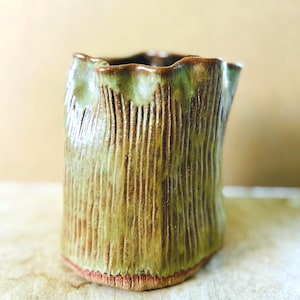 May include: A cylindrical ceramic vessel with a wavy, uneven rim. The exterior features a textured surface with vertical grooves and a variegated glaze in shades of olive green, brown, and hints of reddish-orange at the base of the object.