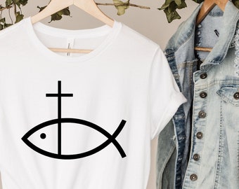 May include: White t-shirt with a black graphic of a fish with a cross on top. The shirt is hanging on a wooden hanger with a denim jacket and a straw hat in the background.