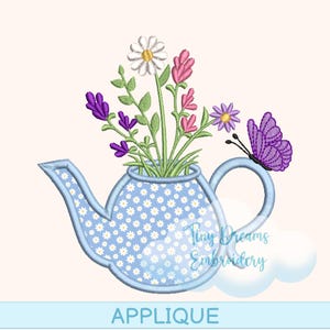 May include: A blue teapot-shaped applique with a floral design, holding a bouquet of flowers in shades of white, pink, purple, and green. A purple butterfly rests on the handle. The word "APPLIQUE" is at the bottom.