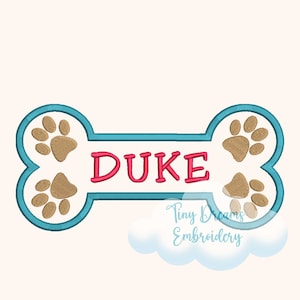 May include: Embroidered dog bone design with the name "DUKE" in pink letters. The bone is white with a teal outline and paw prints. The image also includes the text "Tiny Dreams Embroidery".