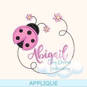 May include: Pink ladybug design with black polka dots and the name "Abigail" in pink script. The design includes pink flowers and a cloud, with the word "APPLIQUE" at the bottom. Tiny Dreams Embroidery is also written in the image.